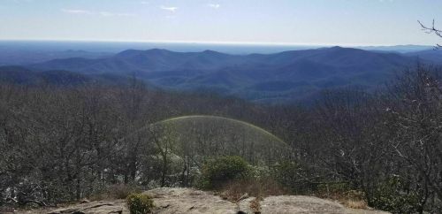 A panoramic view of rolling mountains under a clear sky, with a faint rainbow arching over the landscape.