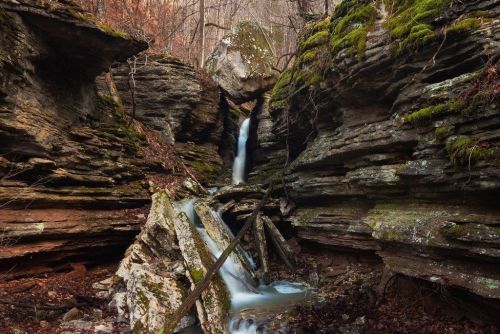 A serene waterfall cascades through rocky terrain, surrounded by moss-covered stones and autumn foliage.