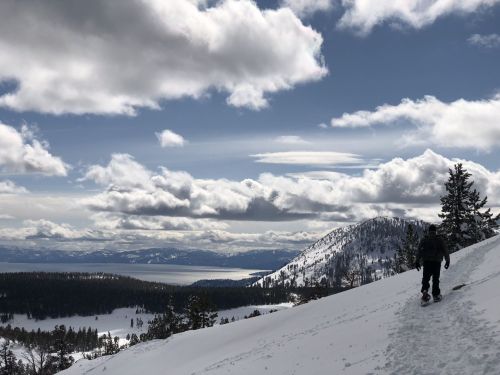 A person snowshoeing on a snowy slope, with mountains and a lake visible under a cloudy sky.