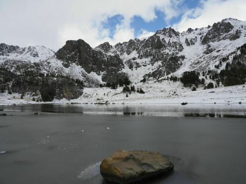 Snow-covered mountains reflect in a calm lake under a partly cloudy sky. A large rock sits in the foreground.