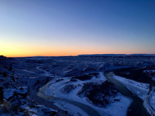 A serene winter landscape at dusk, featuring snow-covered hills and a winding river under a colorful sky.