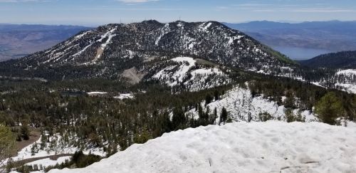 A snowy mountain landscape with green trees, showcasing a rugged terrain and distant hills under a clear blue sky.