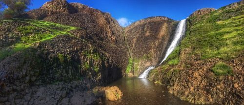 A panoramic view of a waterfall cascading down rocky cliffs, surrounded by lush green vegetation and a serene pool below.