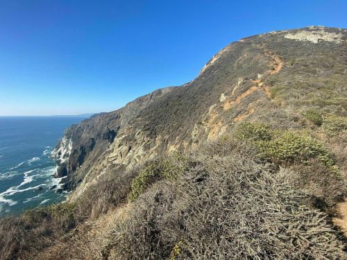 A coastal landscape featuring a rugged cliffside, ocean waves below, and clear blue skies above.