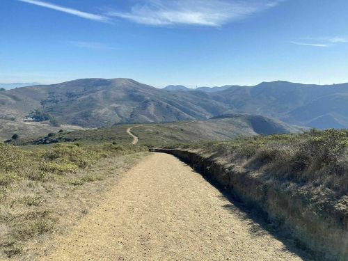 A dirt path winds through grassy hills under a clear blue sky, leading into the distance.