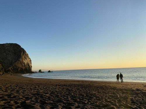 A couple stands on a sandy beach at sunset, with cliffs and calm water in the background.