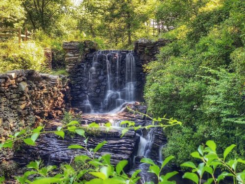 A serene waterfall cascades over rocky ledges, surrounded by lush greenery and trees in a peaceful natural setting.