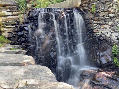 A serene waterfall cascading over rocks, surrounded by greenery and stone walls, creating a peaceful natural scene.