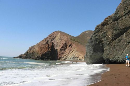 A sandy beach with rocky cliffs and gentle waves under a clear blue sky, with a person walking along the shore.