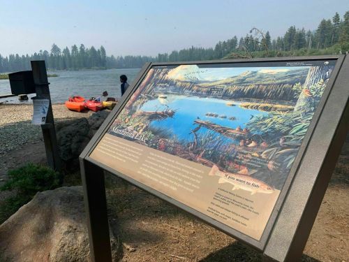 A sign about Mountain Lake near a calm water body, with kayaks on the shore and trees in the background.