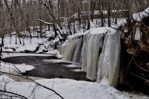 A frozen waterfall cascading into a dark pool, surrounded by snow-covered trees and a winter landscape.