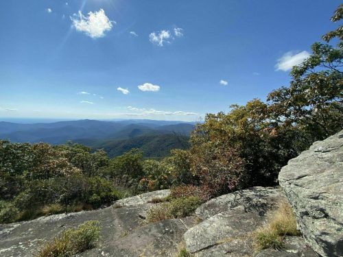 A scenic mountain view with rolling hills, trees, and a clear blue sky dotted with clouds.