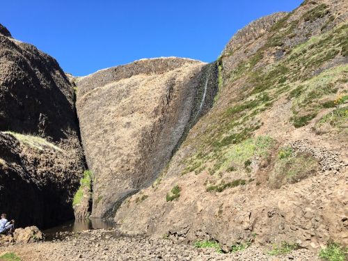 A rocky canyon with a small waterfall cascading down, surrounded by dry, rugged terrain under a clear blue sky.