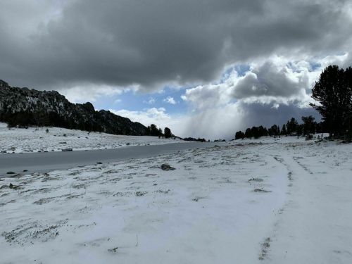 A snowy landscape with rocky hills under a cloudy sky, featuring a river and sparse trees in the distance.