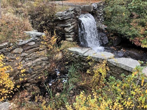 A small waterfall cascades over stone ruins, surrounded by autumn foliage and greenery.