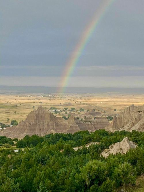 A rainbow arcs over rugged rock formations and green trees in a vast, open landscape.