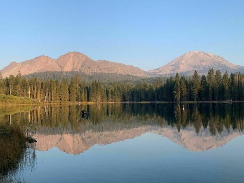 Serene lake reflecting mountains and trees under a clear blue sky, creating a tranquil natural landscape.