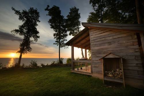 A rustic wooden cabin by the lake at sunset, surrounded by trees and a stack of firewood.