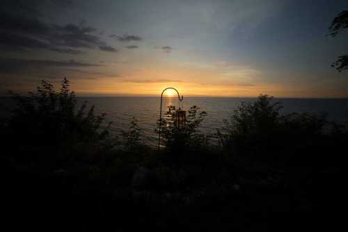 A serene sunset over the ocean, with a wind chime silhouetted against the colorful sky and lush greenery.