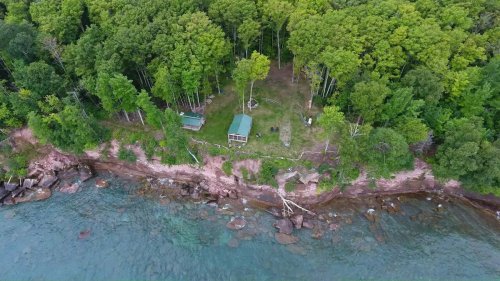 Aerial view of a forested shoreline with a grassy area and a small cabin near the water's edge.