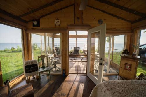 Cozy wooden room with large windows overlooking a lake, featuring a chair, table, and a door leading outside.