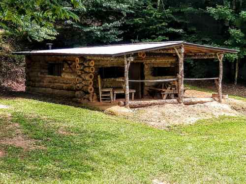 A rustic log cabin with a wooden porch, surrounded by greenery and trees.
