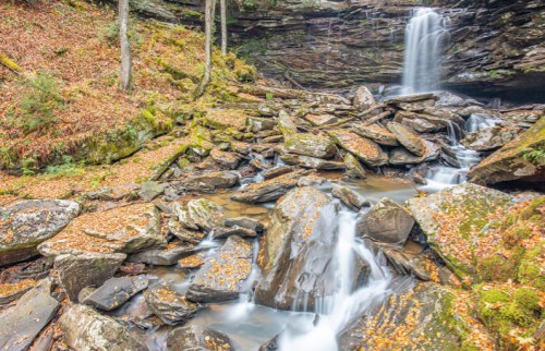 A serene waterfall cascades over rocks, surrounded by autumn foliage and a tranquil stream.