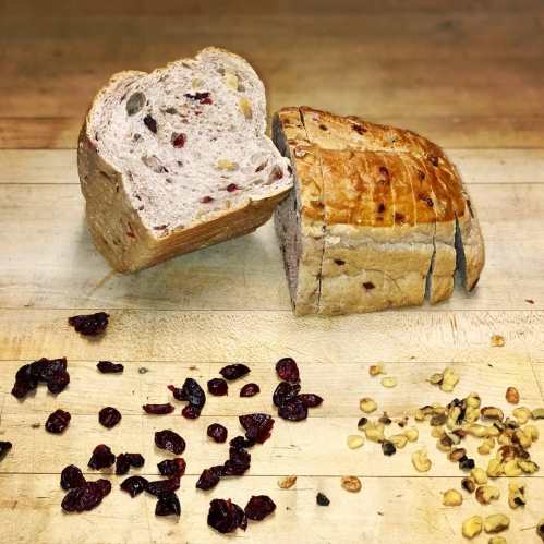Sliced loaf of bread with cranberries and walnuts, surrounded by scattered dried cranberries and walnuts on a wooden surface.