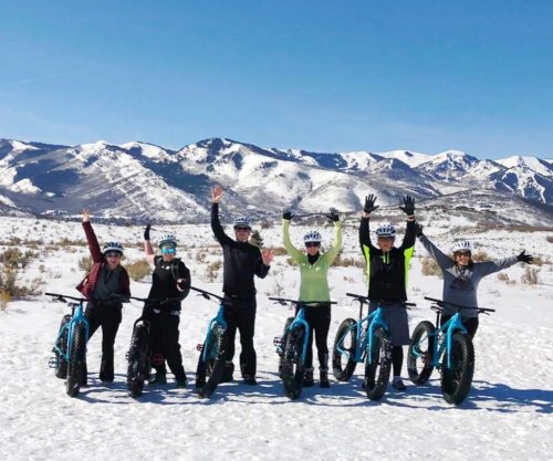 A group of six people on fat bikes in the snow, raising their hands with mountains in the background.