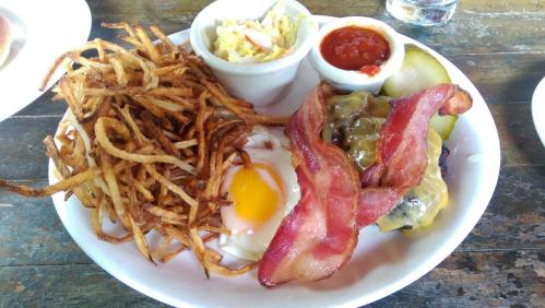 A plate of crispy fries, a sunny-side-up egg, a cheeseburger with bacon, coleslaw, pickles, and dipping sauce.
