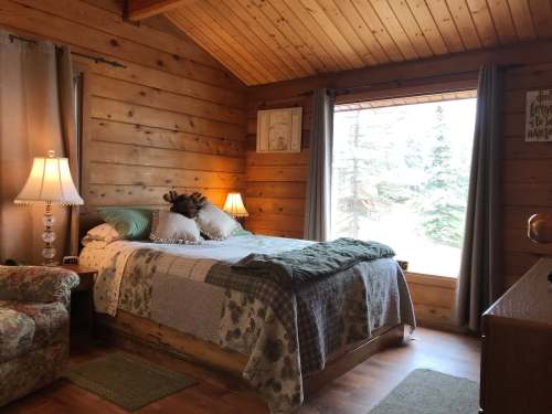 Cozy bedroom with wooden walls, a bed with decorative pillows, a lamp, and a large window overlooking trees.