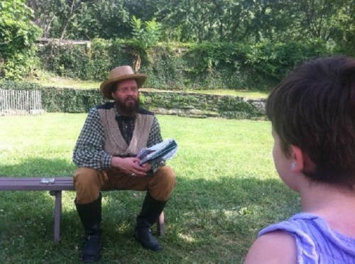 A man in period clothing sits on a bench, reading to a child in a grassy outdoor setting.