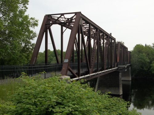 A rusted metal bridge spans a calm river, surrounded by greenery and trees on both sides.