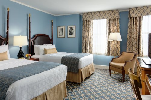 A cozy hotel room featuring two beds, blue walls, patterned curtains, and a desk with a chair.