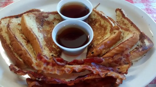 Plate of French toast slices with two cups of syrup and crispy bacon strips on the side.