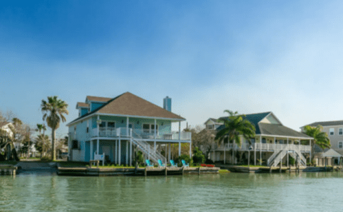 Two waterfront houses with palm trees, blue skies, and a calm waterway in between.