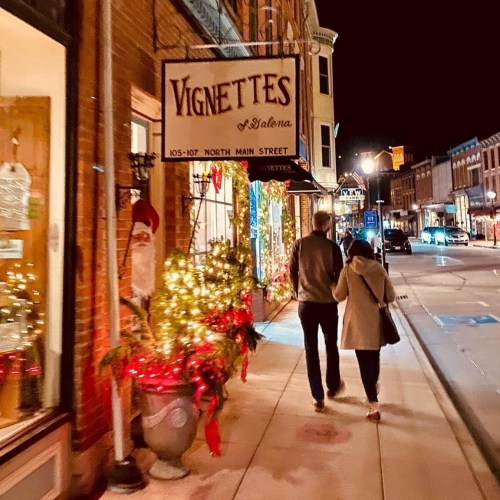 A couple walks down a festive street at night, passing decorated shops and holiday lights.