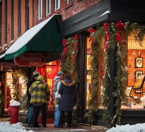 A group of people admire holiday decorations outside a shop adorned with greenery and red bows in a snowy setting.
