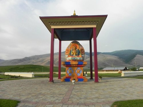 A colorful statue of a deity under a large pavilion, surrounded by green hills and a cloudy sky.