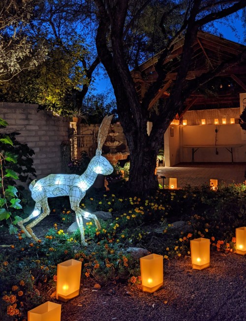 A glowing wireframe rabbit stands among flowers, with lanterns lining a path in a serene evening setting.