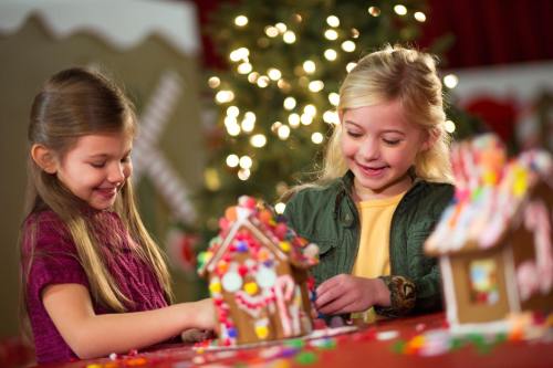 Two young girls joyfully decorating gingerbread houses with colorful candies, with a festive tree in the background.