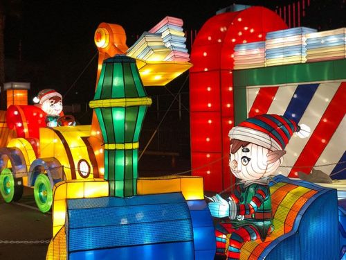 Colorful illuminated display of a train with two cartoonish elves, one driving and one waving, set against a night sky.