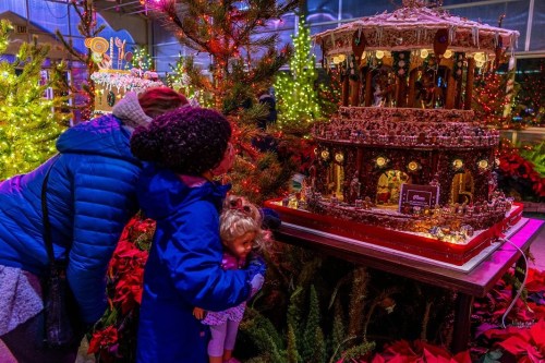 Two people admire a large gingerbread structure decorated with lights, surrounded by festive greenery and holiday decorations.