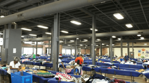 A large indoor space filled with blue bins of clothing, with people sorting through items under bright lights.