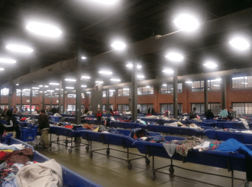 A spacious thrift store with rows of blue bins filled with clothes and shoppers browsing the items.