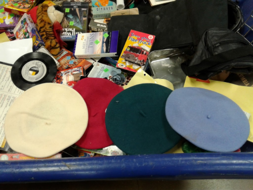 A collection of four berets in different colors (white, red, green, blue) on a cluttered table of various items.