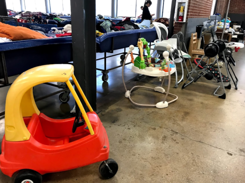 A red toy car and various baby items are lined up in a room with beds and a person in the background.