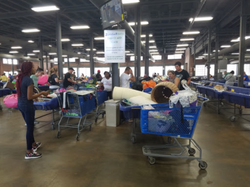 A busy warehouse with people sorting items in shopping carts, surrounded by various goods and a large TV screen.