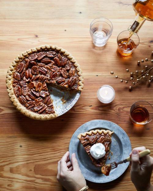 A pecan pie on a wooden table, with a slice on a plate topped with whipped cream and drinks in the background.