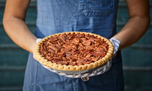 A person holding a freshly baked pecan pie in a pie dish, wearing a blue apron and a checkered cloth.